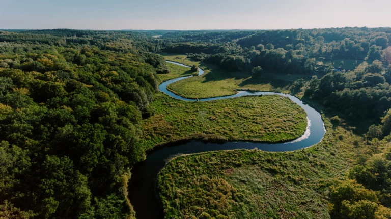 Från ovan syns en å som slingrar sig genom ett böljande landskap av skog och öppna fält i morgonljus.