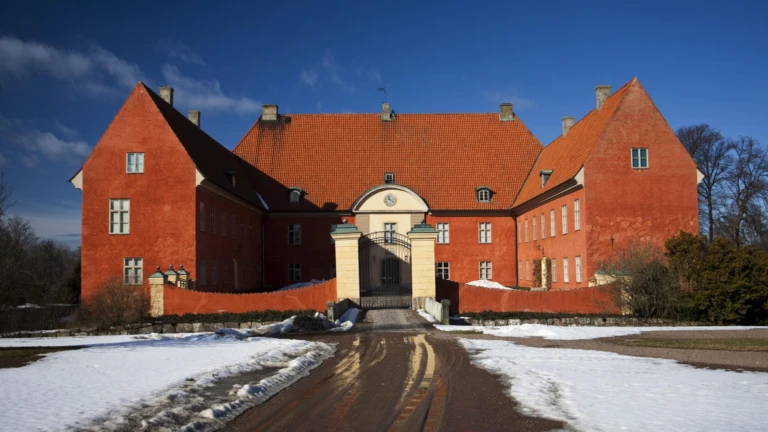 Bild av Krapperups slott i Höganäs kommun, en historisk rödputsad byggnad med höga gavlar och stort tegeltak. Slottet har en ljus portgång i mitten och omges av murar och vinterlandskap med kvarliggande snö. Den klara blå himlen och det varma ljuset framhäver byggnadens färger och arkitektur.