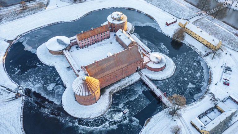 En vintrig flygbild av Landskrona citadell, en historisk fästning byggd i rött tegel och omgiven av en vallgrav täckt av is. Slottet och de intilliggande byggnaderna ligger inbäddade i ett snötäckt landskap med frusna vattenytor och vinterljus som reflekteras i taken.