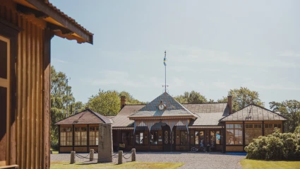 Bild av det militärhistoriska museet i Ljungbyhed Park i Klippans kommun. Den charmiga träbyggnaden har ett randigt tak, klocka och svensk flagga i topp, och omges av grön gräsmatta och träd i sommargrönska. Ljuset och miljön förmedlar en känsla av historia och småskalig idyll.
