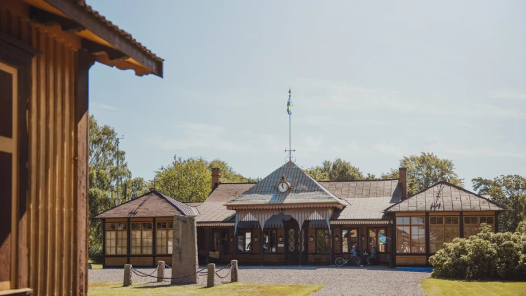 Bild av det militärhistoriska museet i Ljungbyhed Park i Klippans kommun. Den charmiga träbyggnaden har ett randigt tak, klocka och svensk flagga i topp, och omges av grön gräsmatta och träd i sommargrönska. Ljuset och miljön förmedlar en känsla av historia och småskalig idyll.