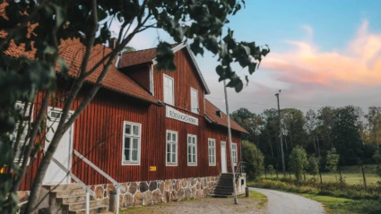 Bild av Röstångagården, en klassisk röd träbyggnad med vita fönster och detaljer, samt tegeltak. Byggnaden ligger i grönskande omgivningar vid en slingrande grusväg, med skog i bakgrunden och en kvällshimmel i mjuka pastellfärger. Miljön förmedlar lugn och landsbygdscharm.