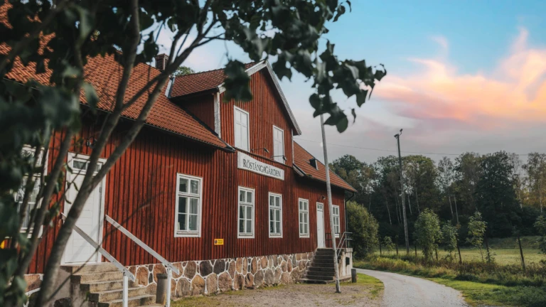Bild av Röstångagården, en klassisk röd träbyggnad med vita fönster och detaljer, samt tegeltak. Byggnaden ligger i grönskande omgivningar vid en slingrande grusväg, med skog i bakgrunden och en kvällshimmel i mjuka pastellfärger. Miljön förmedlar lugn och landsbygdscharm.