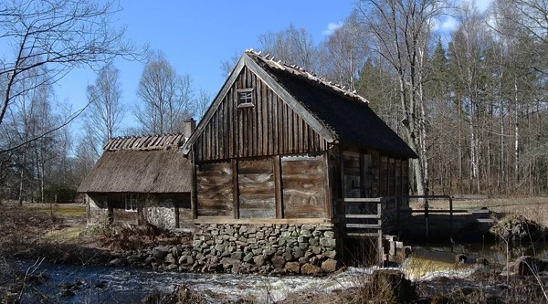 Bild av Rüveröds kvarn i Klippan, en historisk vattenkvarn med timrade väggar och halmtak. Byggnaden står vid en bäck med rinnande vatten och omges av träd i tidig vårgrönska under klarblå himmel.