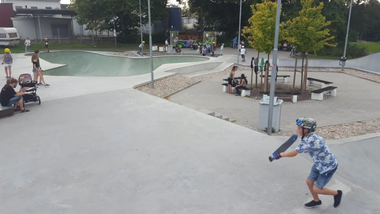 En livlig skatepark där barn och ungdomar åker skateboard och sparkcykel i betongrampens öppna ytor.