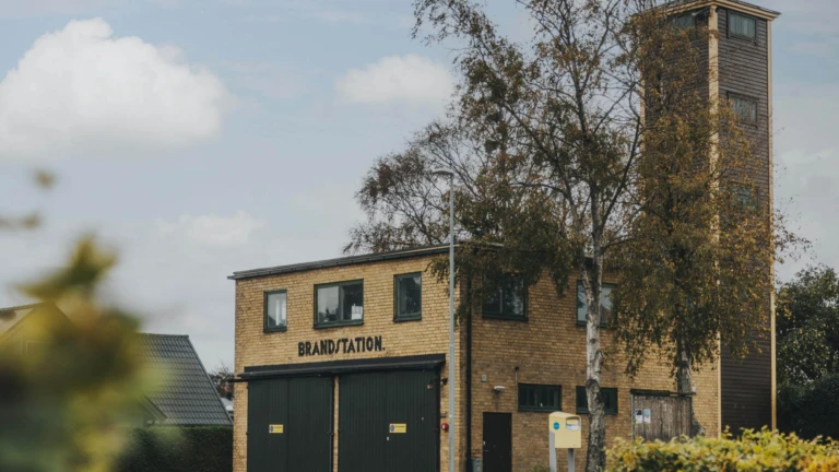 Den lilla brandstationen i Tågarp med sin karakteristiska tornbyggnad står omgiven av träd och bostadshus.