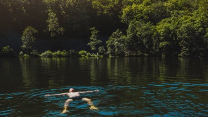 En badande person ligger avslappnat i det klara vattnet i Odensjön, omgiven av täta gröna träd som speglar sig i sjön.