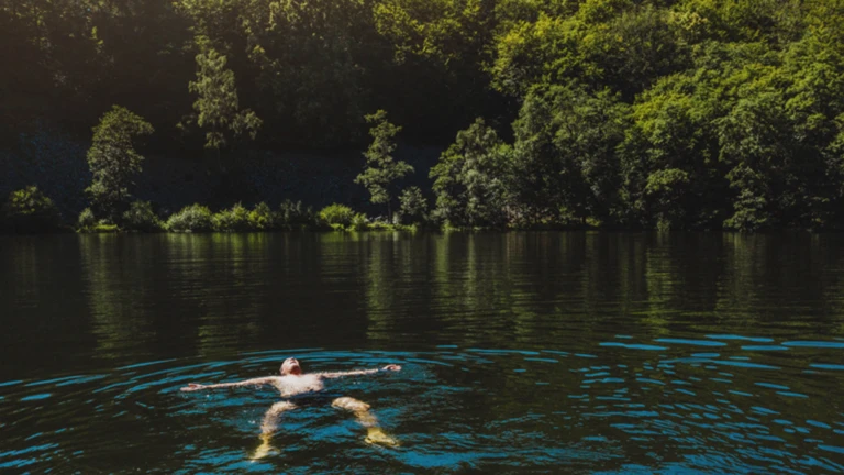 En badande person ligger avslappnat i det klara vattnet i Odensjön, omgiven av täta gröna träd som speglar sig i sjön.