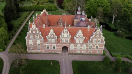 Bild av Vrams Gunnarstorps slott i nordvästra Skåne, ett praktfullt renässansslott byggt i rött tegel med rikt dekorerade gavlar och rött tegeltak. Slottet ligger omgivet av välskötta trädgårdar, grusgångar och grönskande parkmiljö. Fotot är taget från ovan och visar byggnadens symmetri och natursköna omgivning.