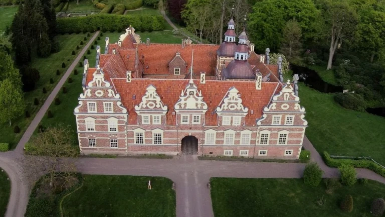 Bild av Vrams Gunnarstorps slott i nordvästra Skåne, ett praktfullt renässansslott byggt i rött tegel med rikt dekorerade gavlar och rött tegeltak. Slottet ligger omgivet av välskötta trädgårdar, grusgångar och grönskande parkmiljö. Fotot är taget från ovan och visar byggnadens symmetri och natursköna omgivning.