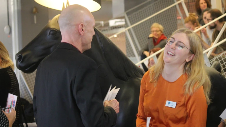 Avslappnat samtal under mingel där två deltagare skrattar tillsammans, med andra i bakgrunden.