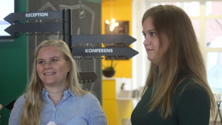 Två deltagare står och samtalar i en ljus lokal med tydliga riktningsskyltar som visar vägen till reception och konferens.