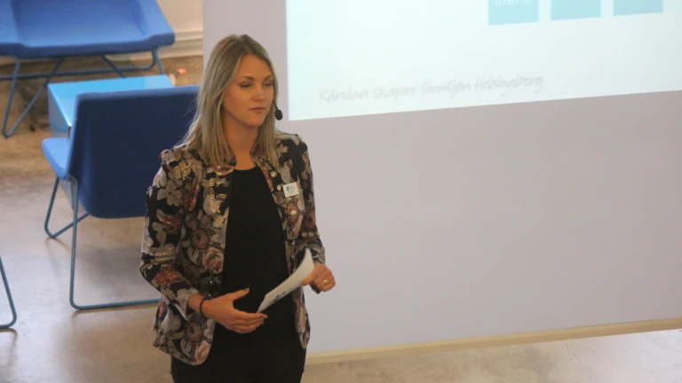 En föreläsare står på scen och håller presentation inför publik i en modern lokal.