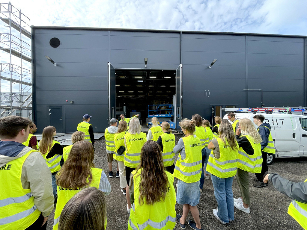 En stor grupp besökare i gula varselvästar framför en fabriksbyggnad med öppna portar