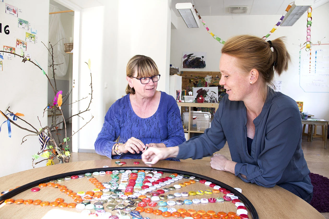 Två pedagoger sitter vid ett stort bord och bygger mandala med kapsyler