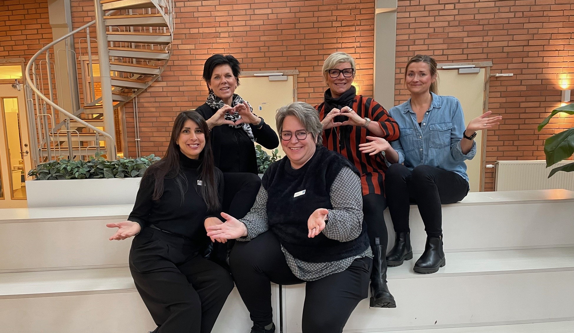 Fem kvinnor som ser glada och välkomnande ut sitter på en inomhustrappa.