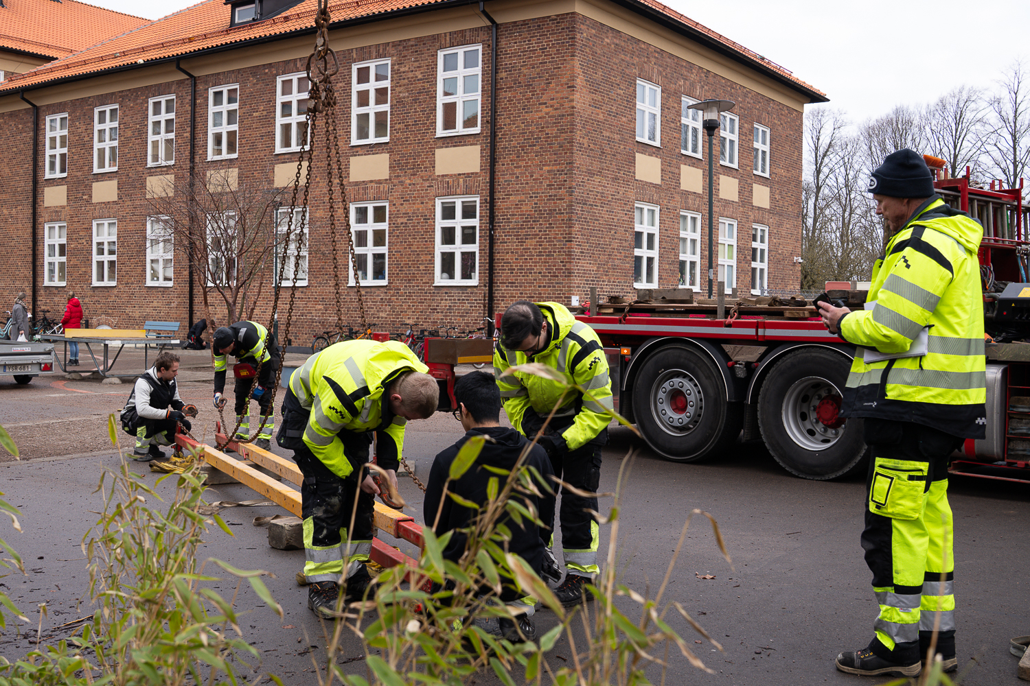 Elever och lärare i byggarbetskläder är aktiva på en skolgård med lastbil med kran