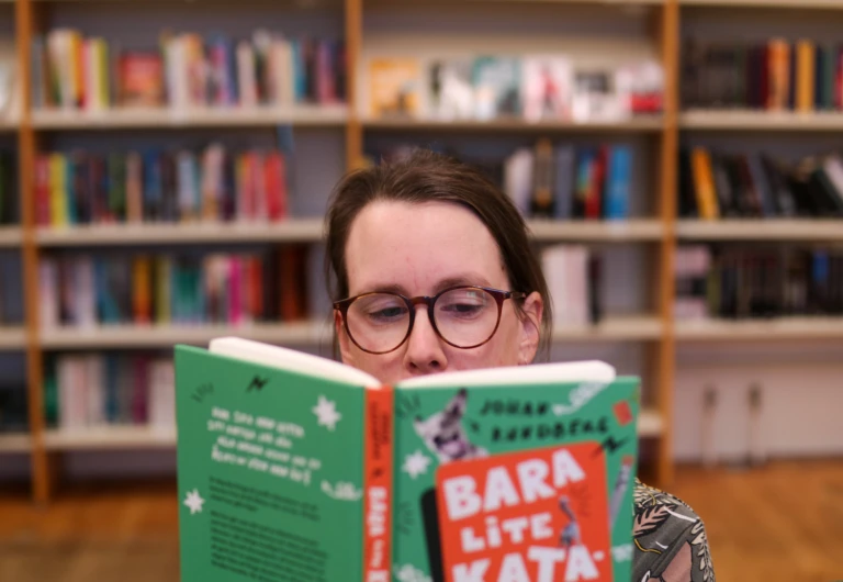 En kvinna sitter framför en stor bokhylla och håller en uppslagen bok framför sig.