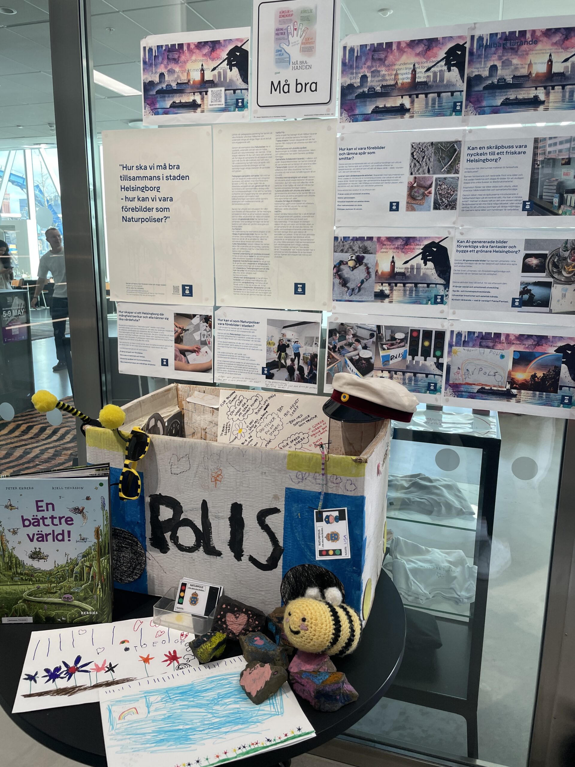 En fysisk utställningsmonter med en kartong märkt ”POLIS”, handmålade teckningar på blommor, stenar med hjärtan och en virkad humla. Bakom står informationsskyltar om projektet Naturpoliser.