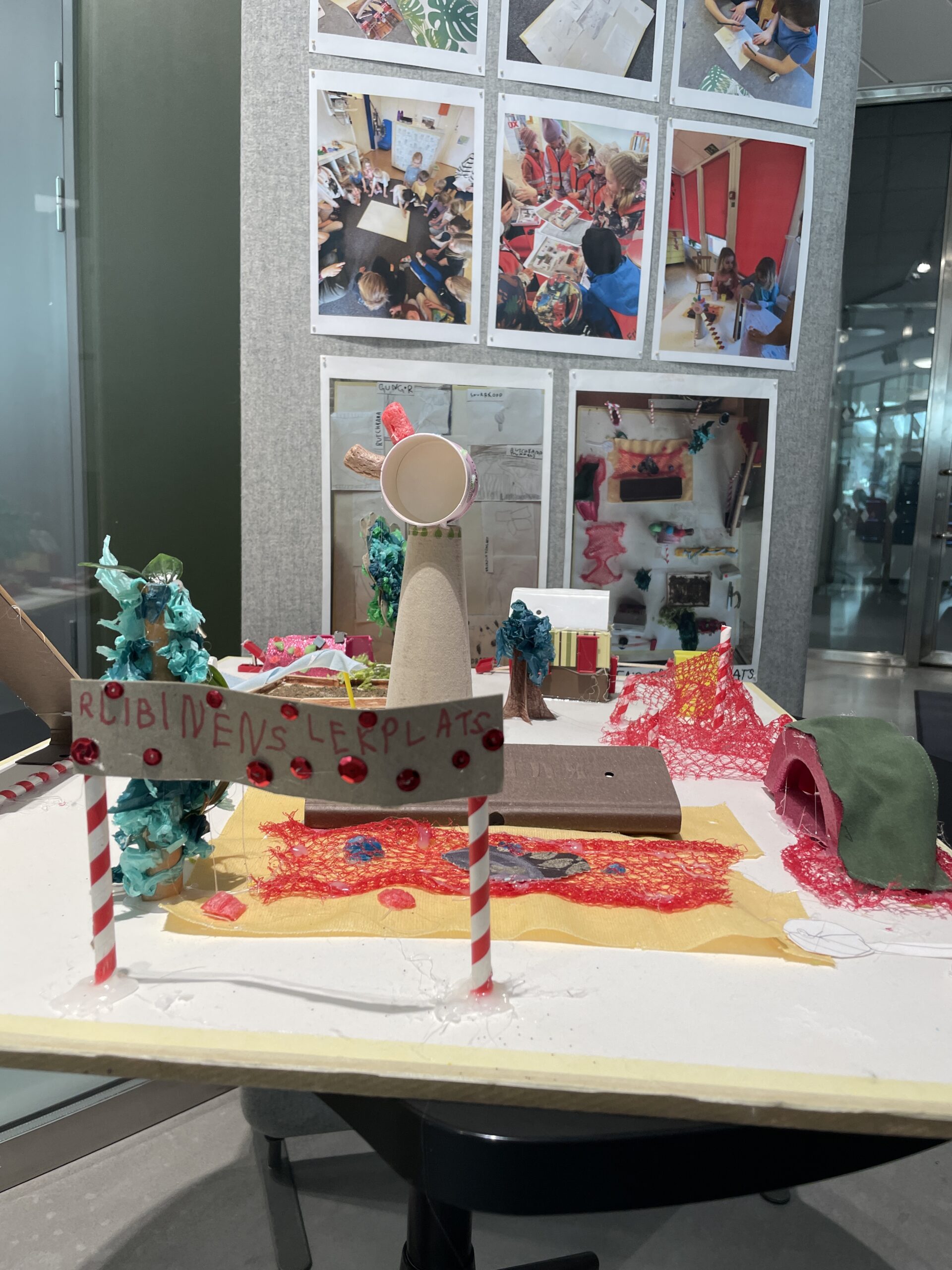 En kreativ modell av en lekplats byggd av återvinningsmaterial som pappmuggar, nät, piprensare och små glittriga detaljer. I bakgrunden syns foton på barn som arbetar med projektet.