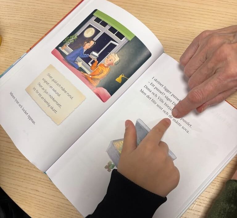Ett barn och en vuxen pekar på en text i en uppslagen bok, vilket visar ett engagerande exempel på gemensam läsning i projektet Familjen läser.