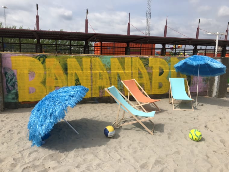 Bilden visar en bit strand med tre solstolar, två parasoller och två bollar. På en vägg bakom stranden har namnet "Banana Bay" målats.