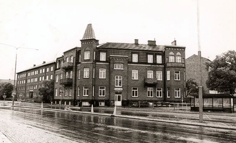 Svartvitt foto med trevånings hörnhus med torn.