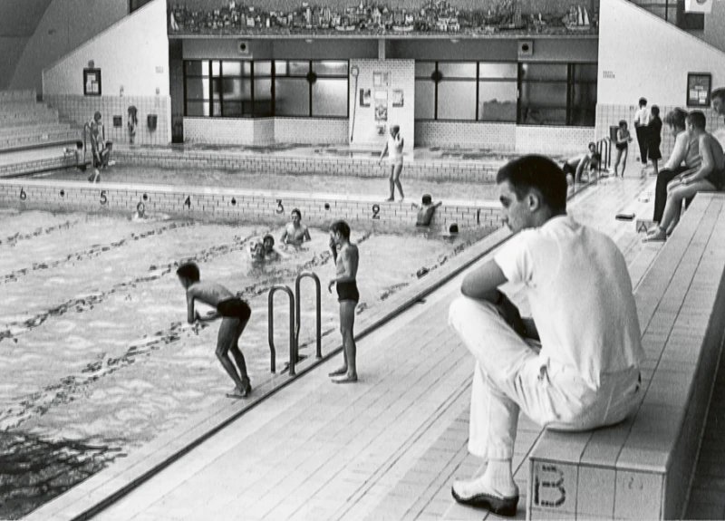 Interiör badhus med simmande barn och en badvakt sittande på läktaren. 