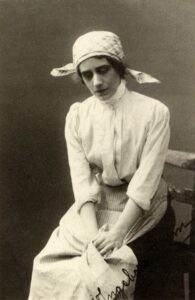 Svartvitt porträtt av sittande kvinna i huckle, blus och förkläde. Bilden föreställer en skådespelare som gestaltar rollen Ingeborg Holm,