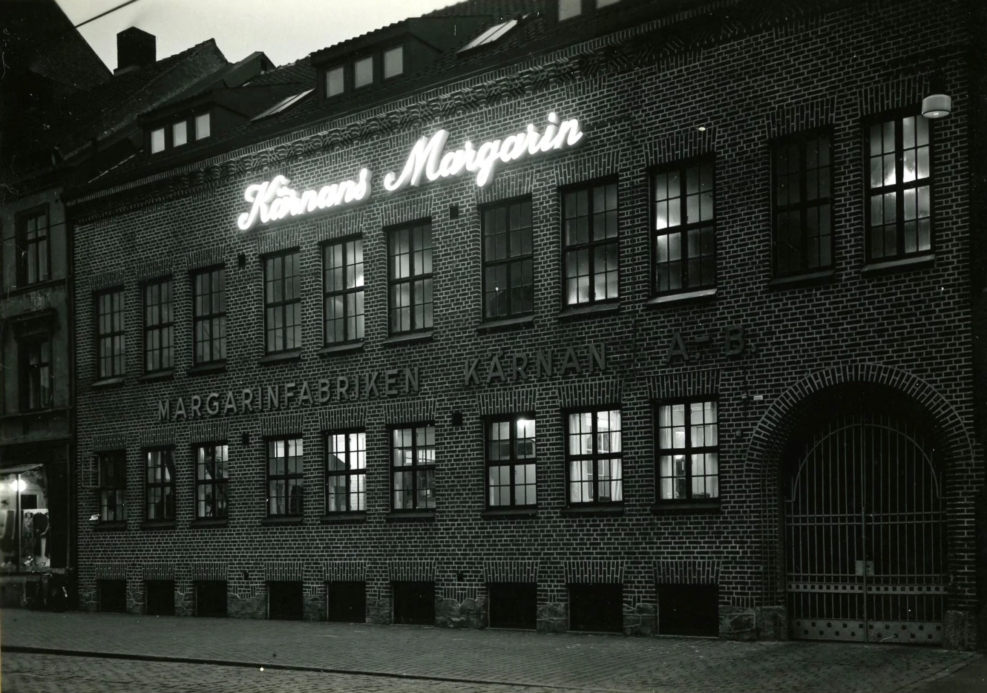 Margarinfabriken Kärnan ABs upplysta fasad kvällstid. Adress: Södergatan 82. Fotograf: Tönnessen. Fotot tillhör Jan Sendelius.