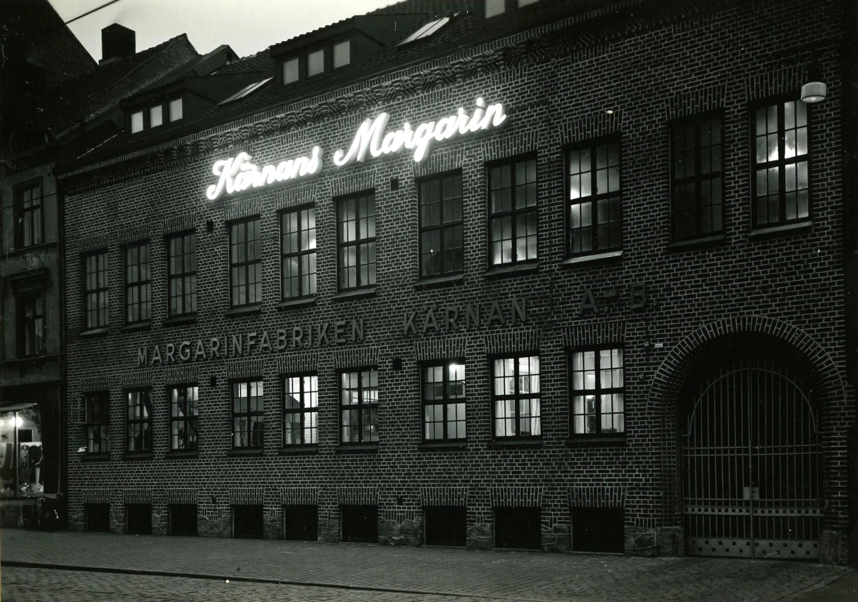 Margarinfabriken Kärnan ABs upplysta fasad kvällstid. Adress: Södergatan 82. Fotograf: Tönnessen. Fotot tillhör Jan Sendelius.