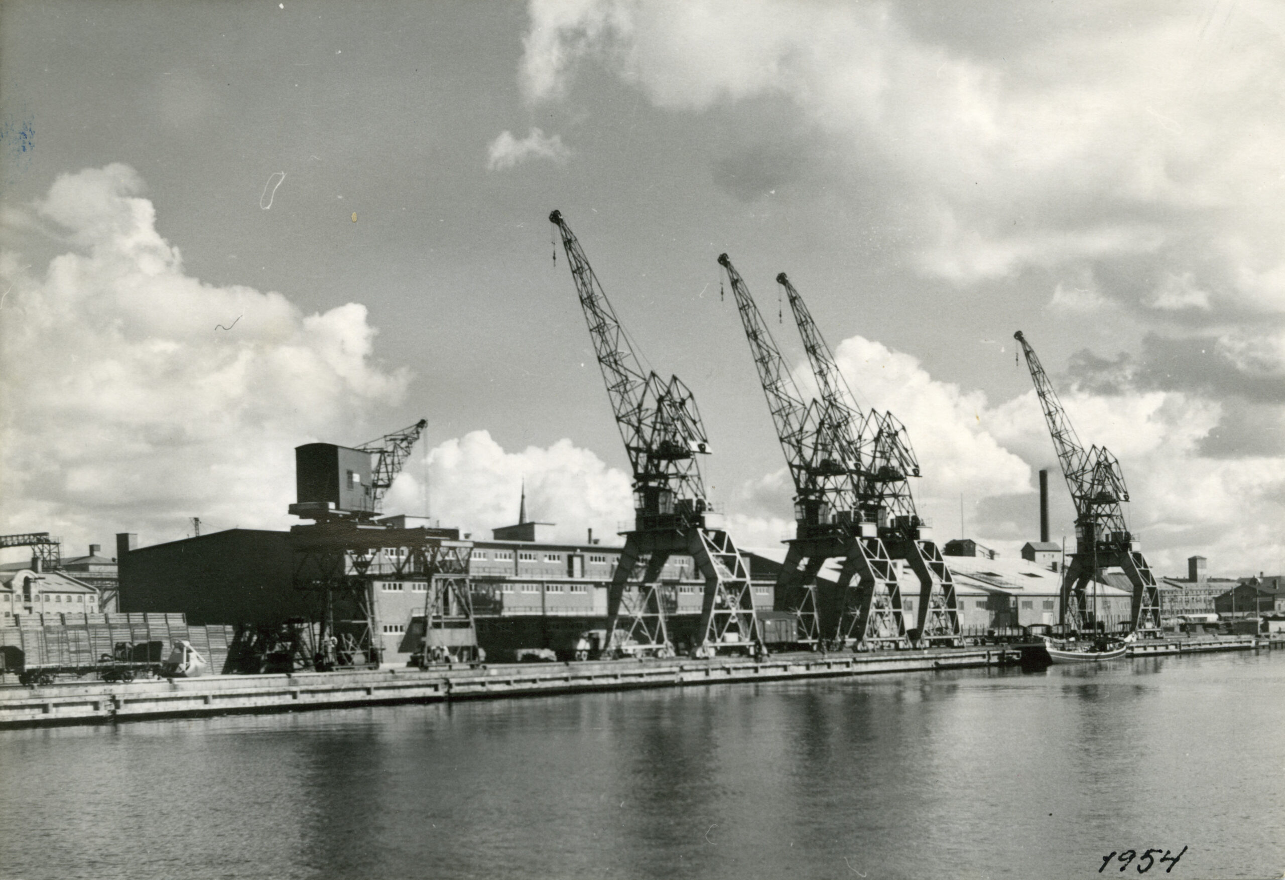 Svartvitt foto på hamnen taget från vattnet. Stora kranar dominerar bilden.