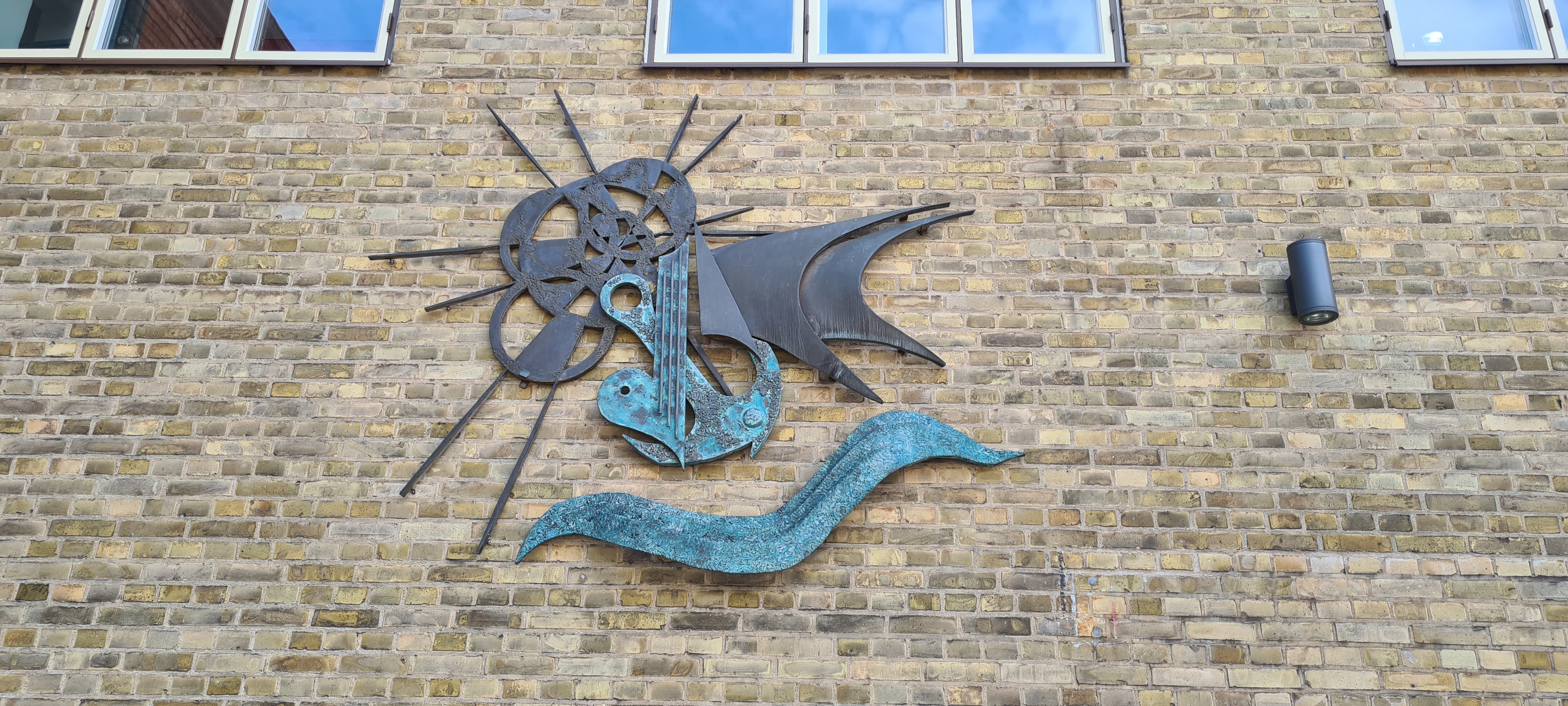 Konstverk i metall på tegelvägg. Mjuka former som bildar sol, segel, vatten ankare och hjärta.