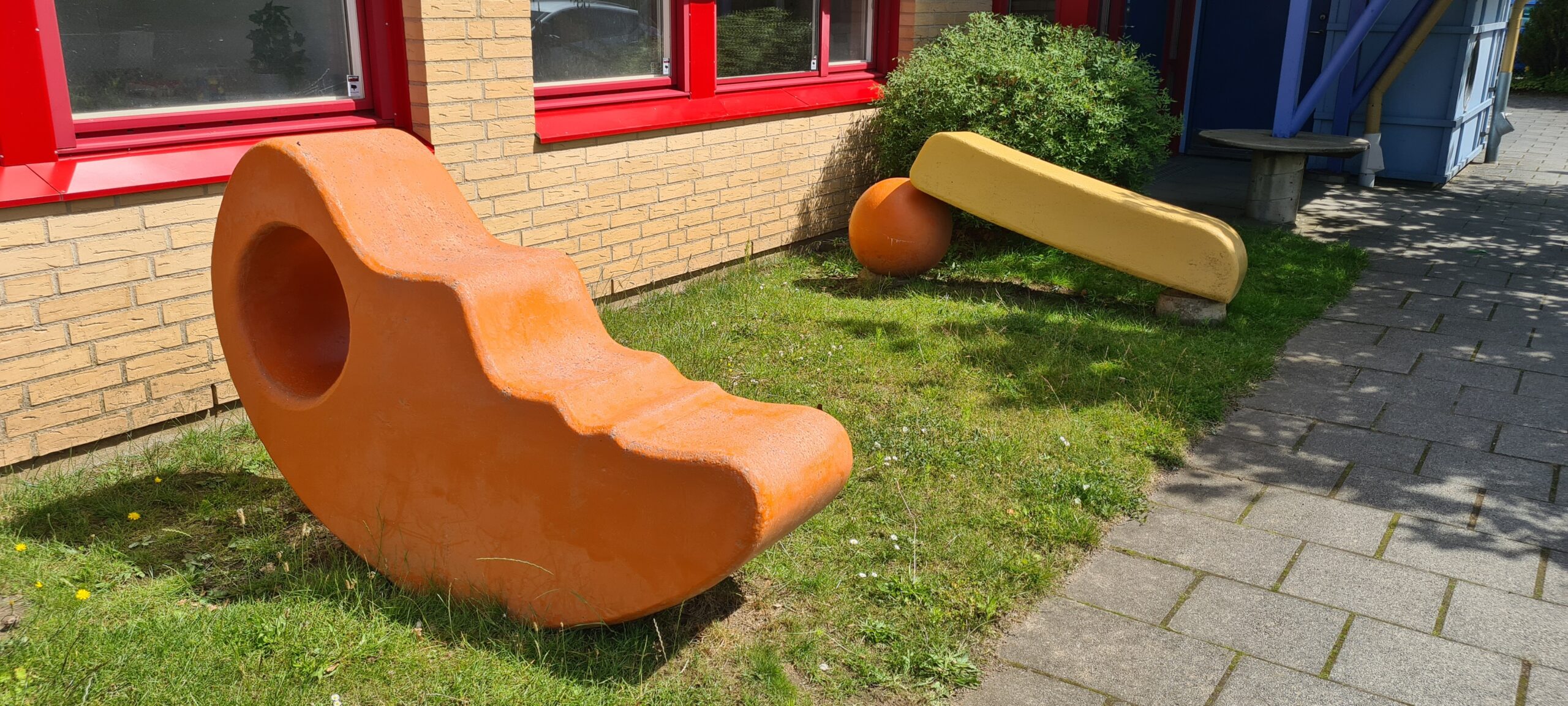 På gräsmatta framför hus, låga betongskulpturer i gul och orange. Föreställer en måne och en solstrimma.