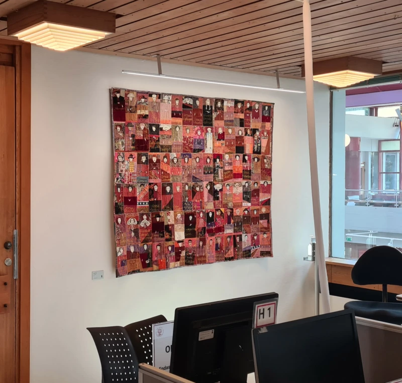 Textiltavla på vägg i rödaktiga färger med rader av kvinnor. 