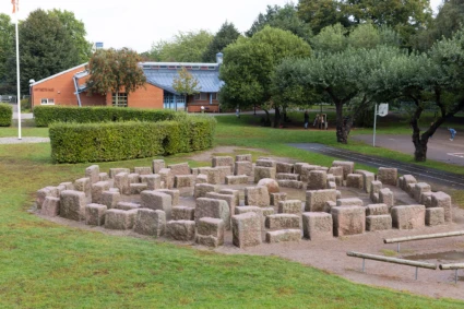 Utomhusbild med gräsplan och skolhus i bakgrunden. I förgrunden en labyrint av stenbumlingar.