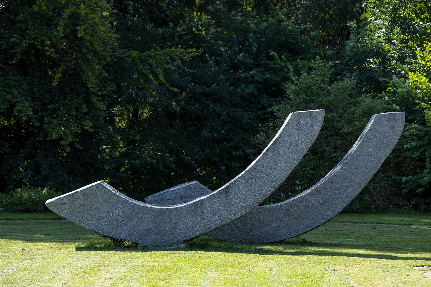 Utomhusbild med skog, gräsmatta och stor granitskulptur i form av två vågor.
