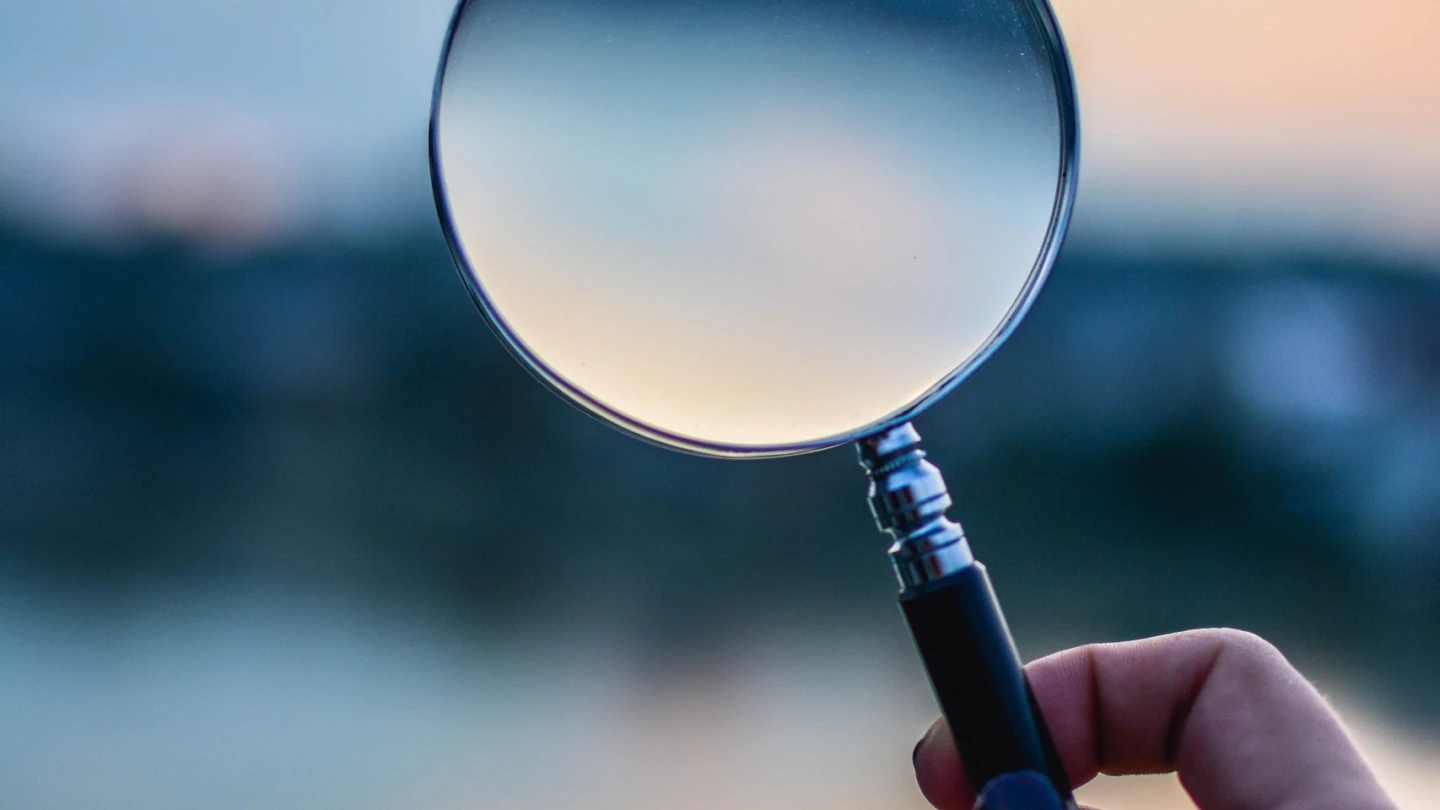 En hand håller upp ett förstoringsglas mot en suddig bakgrund, troligen hav och himmel