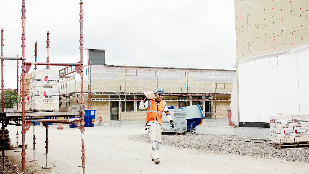 En byggarbetsplats. En byggarbetare bär på några plankor iklädd brandgul väst.