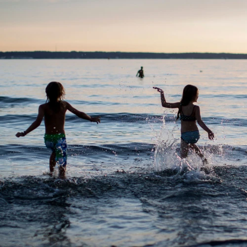 Siluetter av tre personer (bakifrån). De springer ut i havet. Det är sommar och varmt och solen håller på att gå ner.