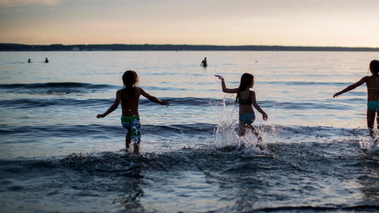 Siluetter av tre personer (bakifrån). De springer ut i havet. Det är sommar och varmt och solen håller på att gå ner.