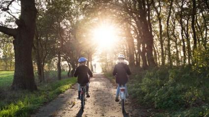 Två barn cyklar på en grusstig omgiven av träd. Solen håller på att gå ner.