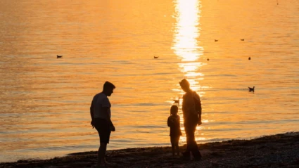 Tre personer står i vattenbrynet på en strand, medan relfektionerna från en solnedgång syns syns i vattenytan.