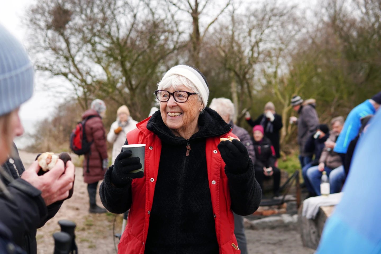 En äldre kvinna står utomhus med en kopp kaffe i handen och pratar med människor.