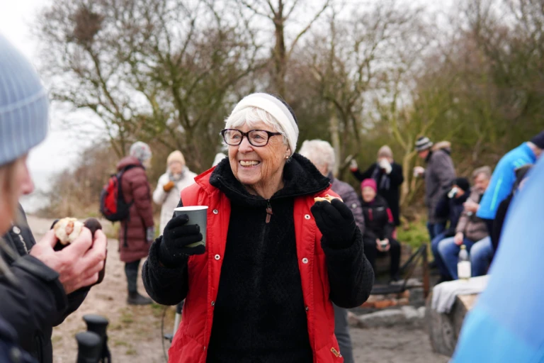 En äldre kvinna står utomhus med en kopp kaffe i handen och pratar med människor.