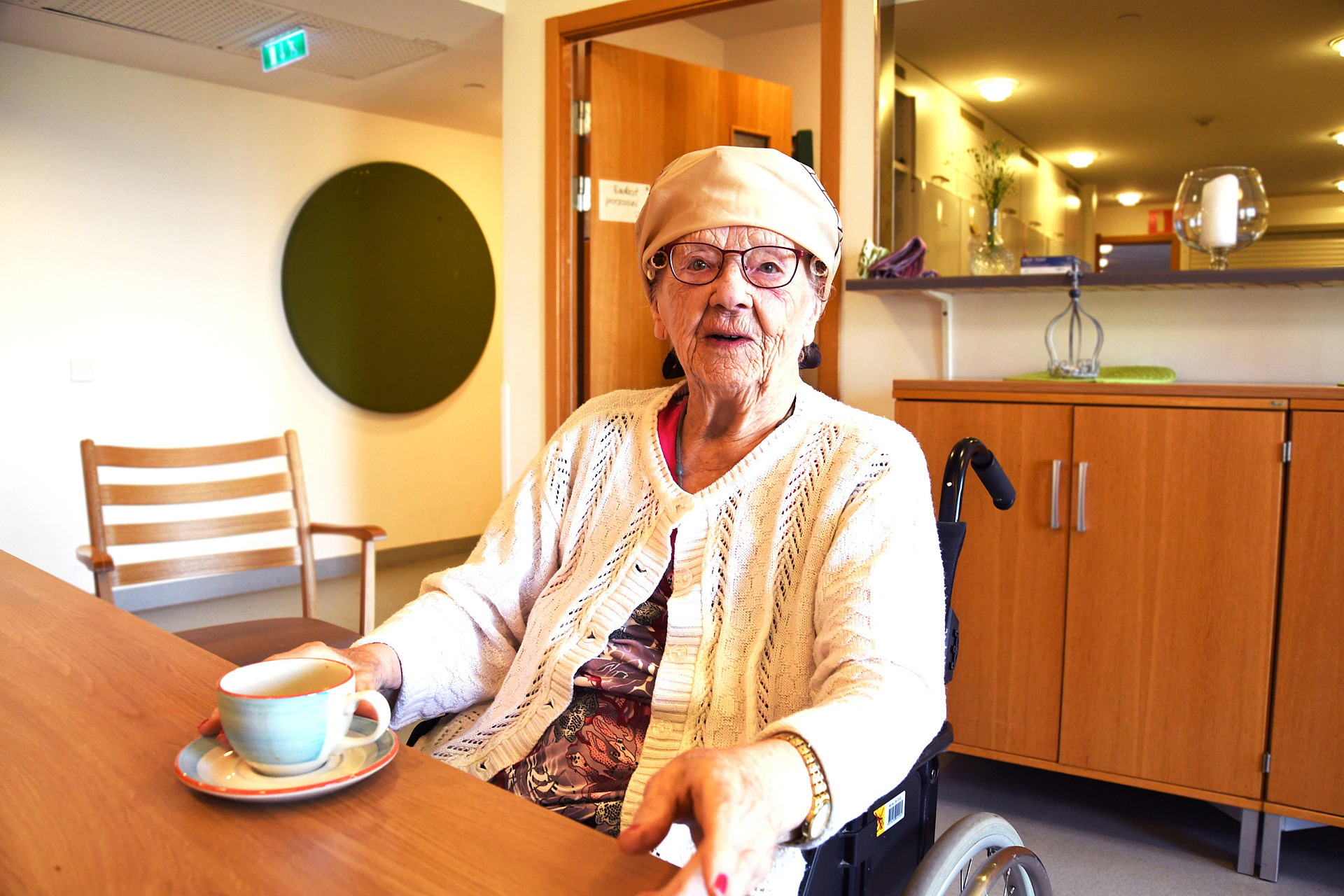 En äldre dam dricker kaffe vid ett bord och tittar in i kameran