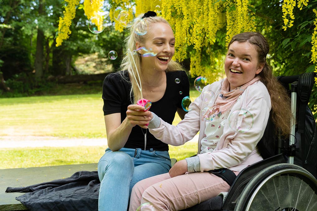 En kvinna ler och tittar på en tjej i rullstol som blåser såpbubblor.
