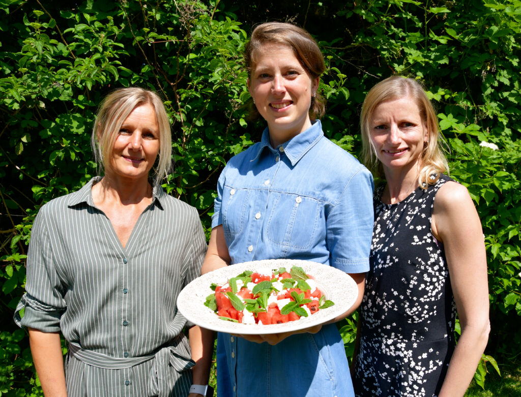 Tre kvinnor står framför grönska, Kvinnan i mitten håller fram en tallrik med melon- och fetaostsallad.
