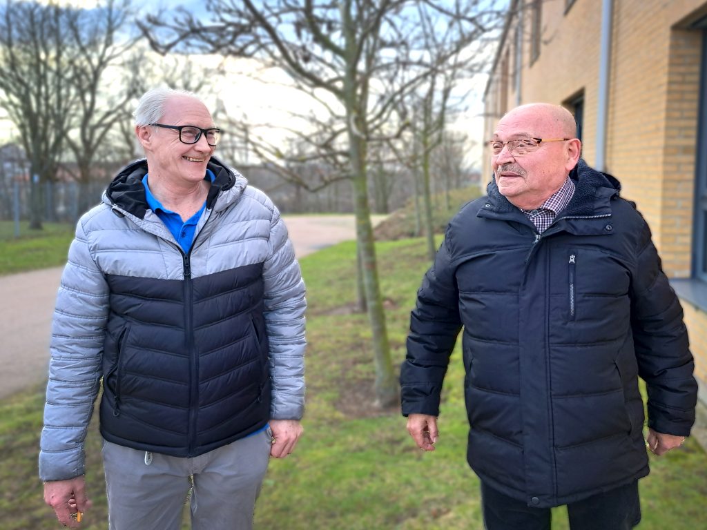 Två män står utomhus. Mannen till vänster har en täckjacka i två färger och huva. Mannen till höger har en mörkblå täckparkas. Båda har glasögon och skrattar.