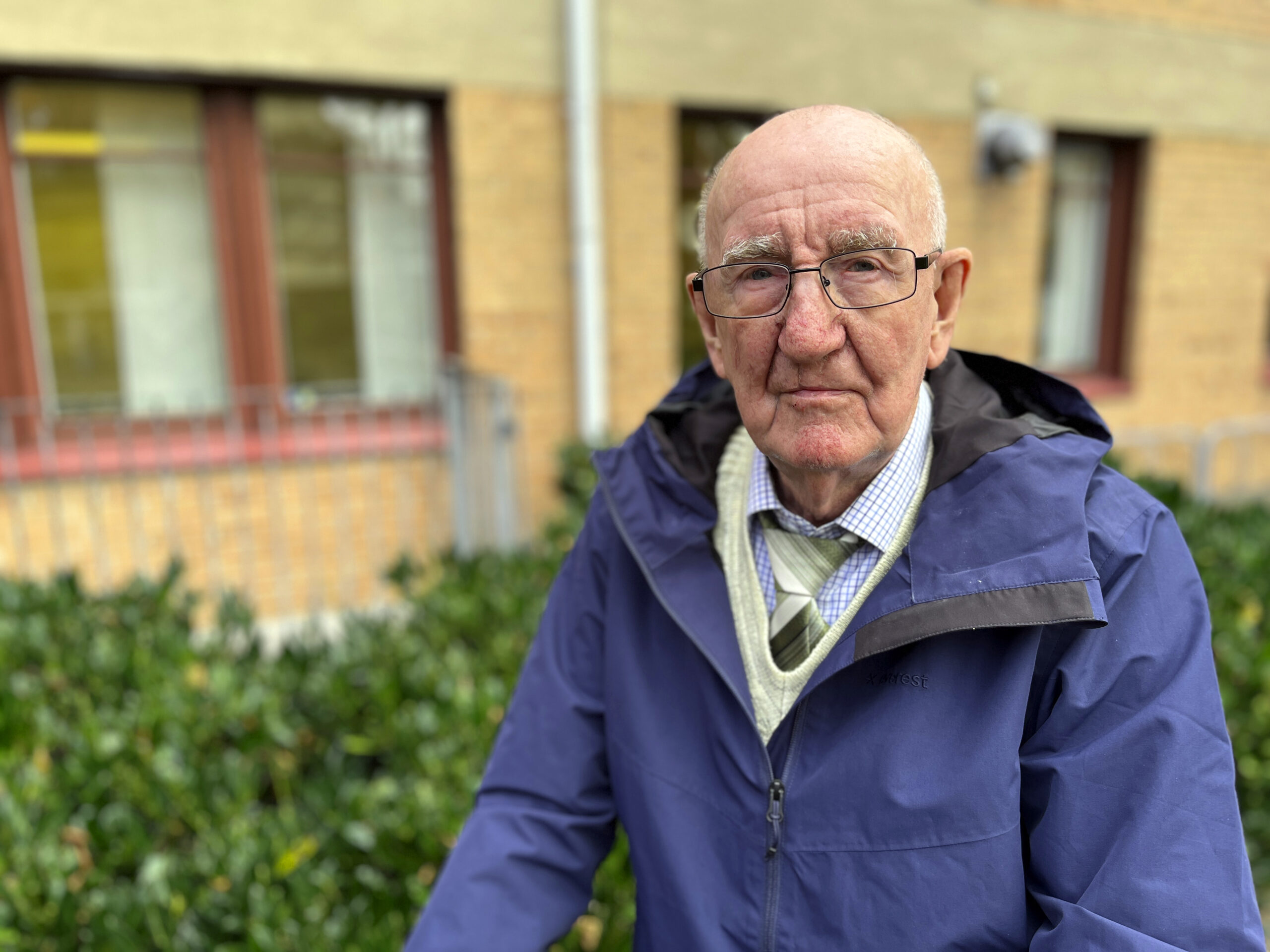 Äldre man med rollator står utomhus framför ett buskage och en tegelbyggnad 